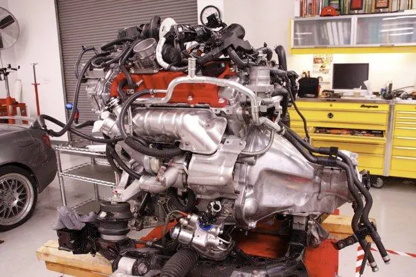 A performance engine assembly on a stand in a tuning shop, featuring red intake manifolds and silver piping.