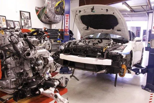 A white Nissan GT-R undergoing an engine swap or build in a performance tuning shop, with the engine removed and on a stand.