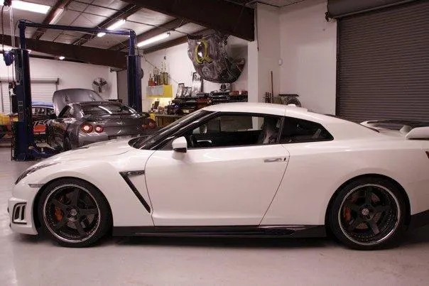 A white Nissan GT-R with custom wheels parked in a performance tuning shop with another car on a lift in the background.
