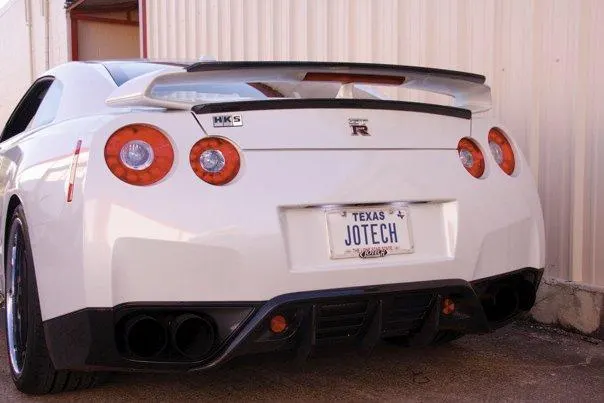 Rear view of a white Nissan GT-R R35 with HKS exhaust, aftermarket wing, and Texas JOTECH license plate.