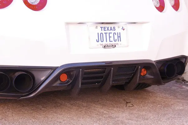 Close-up of a white Nissan GT-R's rear, showcasing a carbon fiber diffuser, aftermarket exhaust tips, and a Jotech Texas license plate.