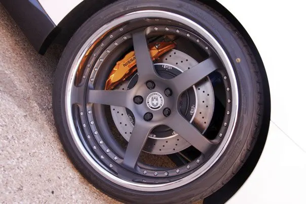 A close-up of a custom matte grey wheel with a polished lip, gold Nissan brake caliper, and drilled rotor on a performance car.