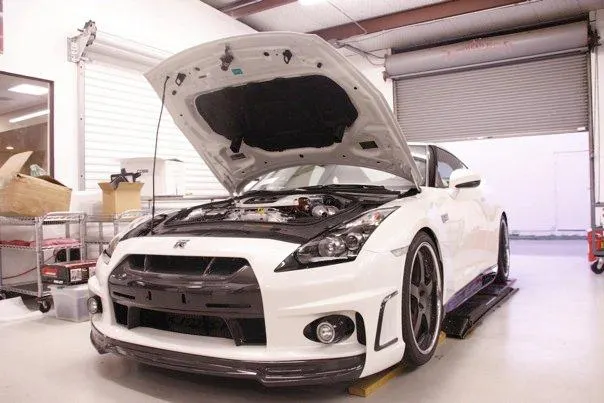 A white Nissan GT-R with its hood open, revealing the engine bay and turbochargers, inside a performance tuning shop.