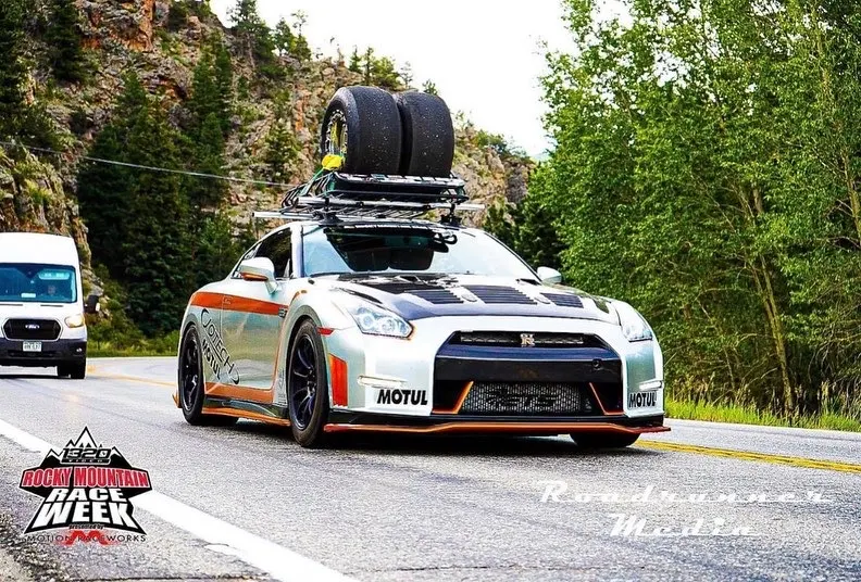 A silver Nissan GT-R with a roof rack carrying race tires drives on a scenic mountain road during Rocky Mountain Race Week.