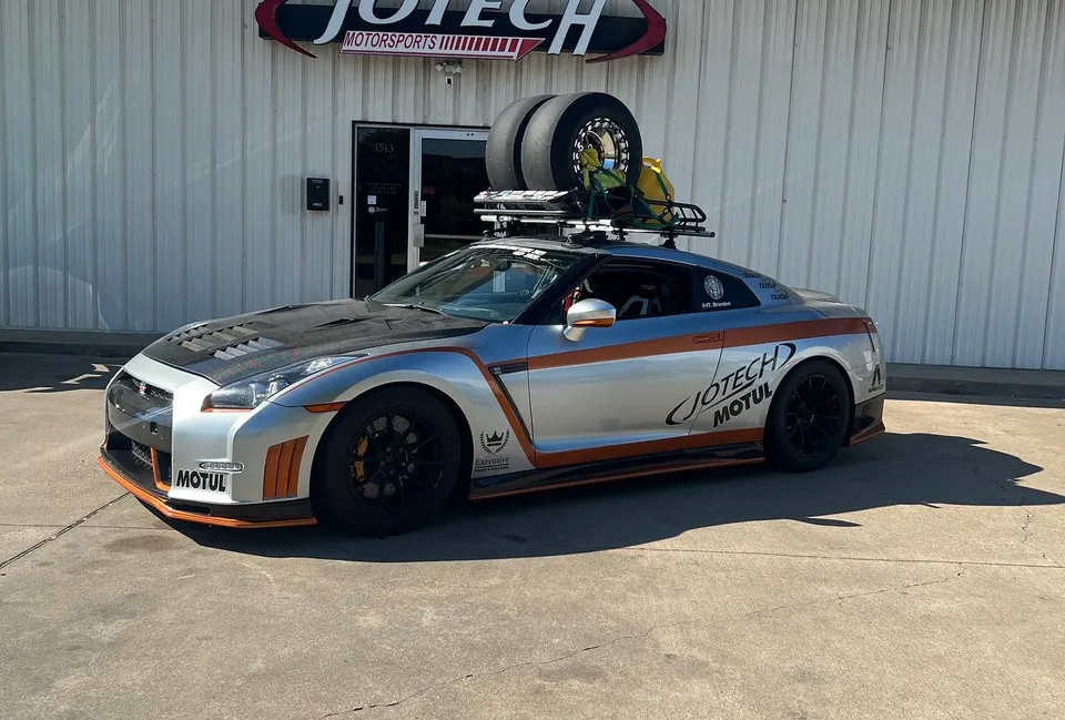 Silver and orange Nissan GT-R R35 with a carbon fiber hood and roof rack carrying race tires parked outside Jotech Motorsports.
