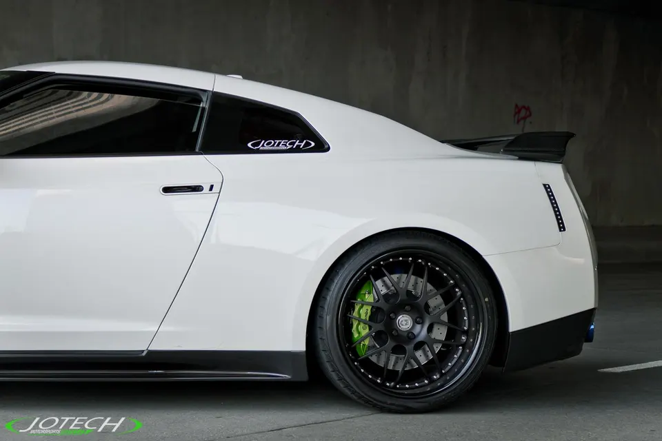 A white Nissan GT-R with aftermarket black wheels, green brake calipers, and a carbon fiber spoiler.