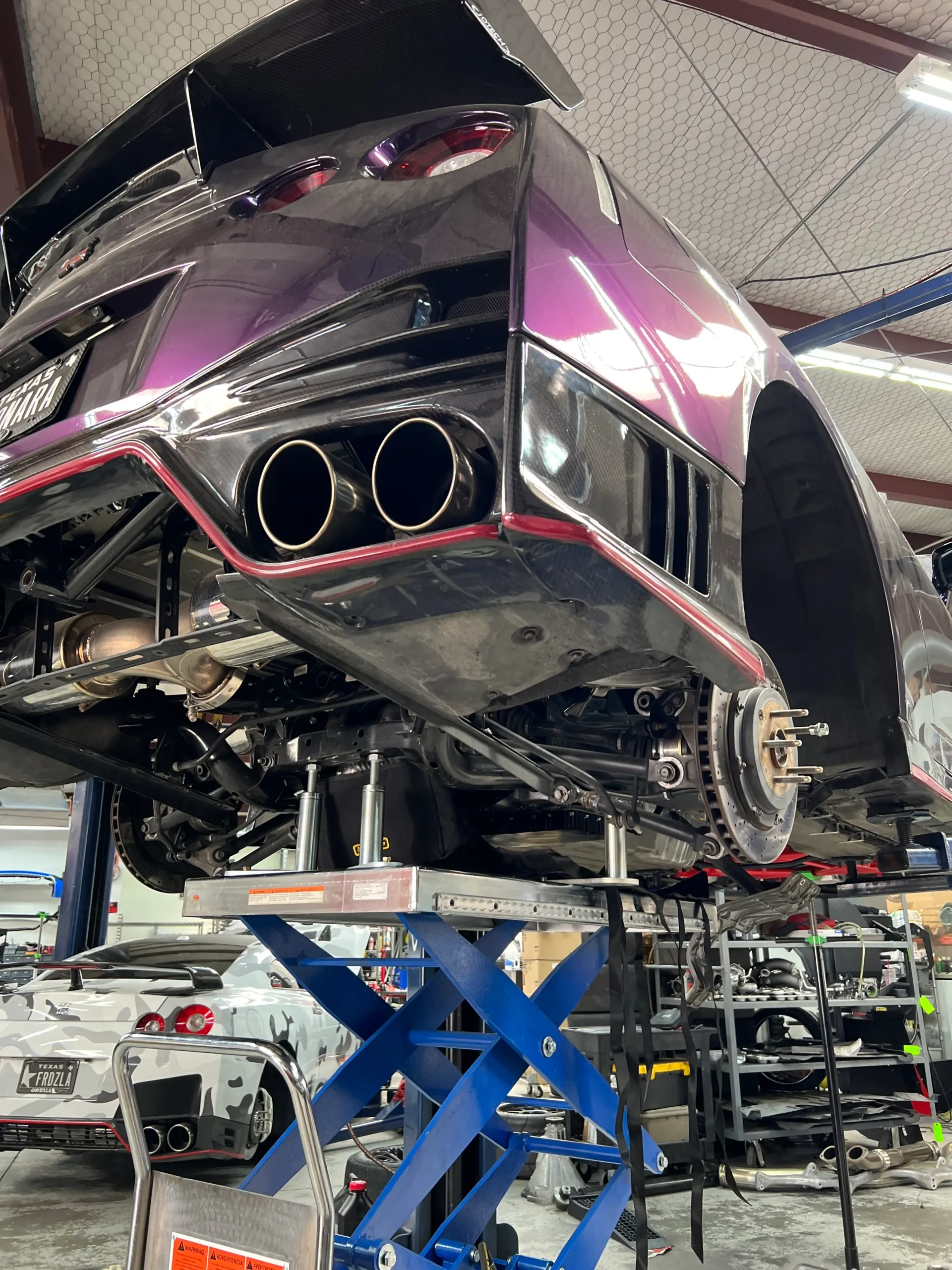 Purple Nissan GT-R on a lift in a performance shop, showcasing its aftermarket exhaust and carbon fiber rear diffuser.