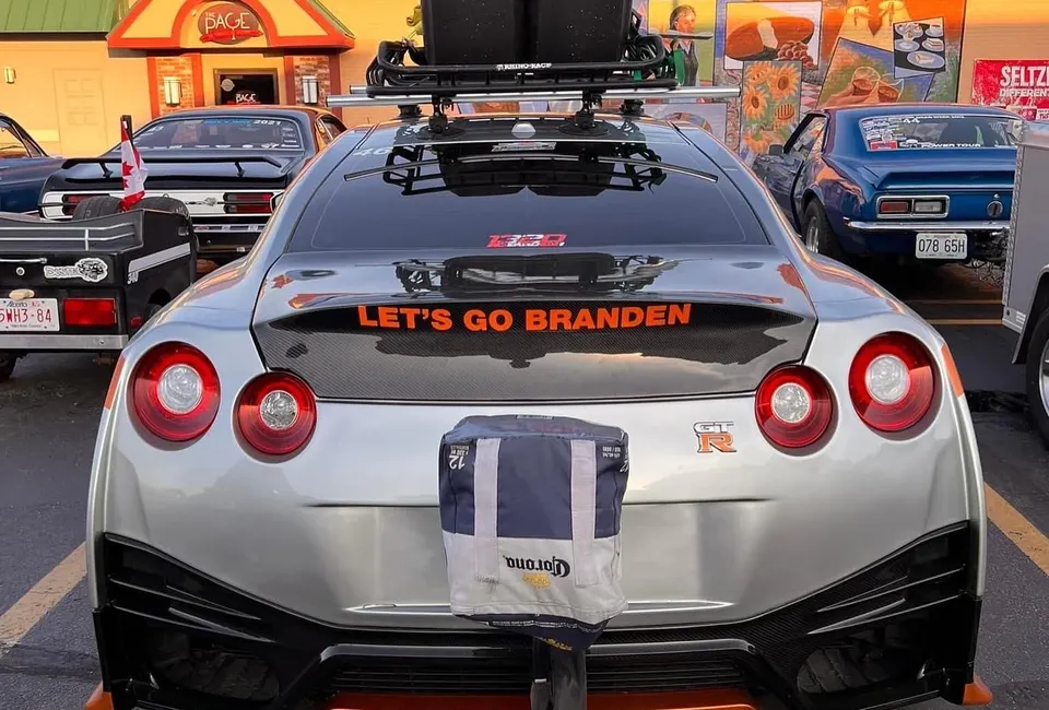 Rear view of a silver Nissan GT-R with a roof rack and a cooler attached, parked in a lot with other cars.
