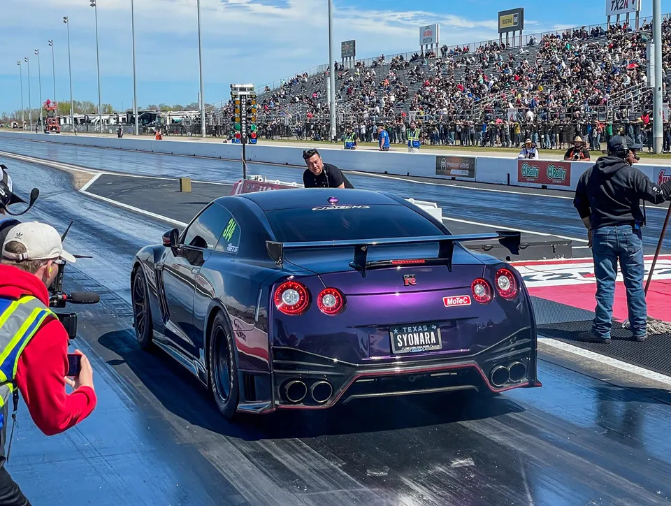 A purple Nissan GT-R on a drag strip with spectators in the stands, preparing for a race.