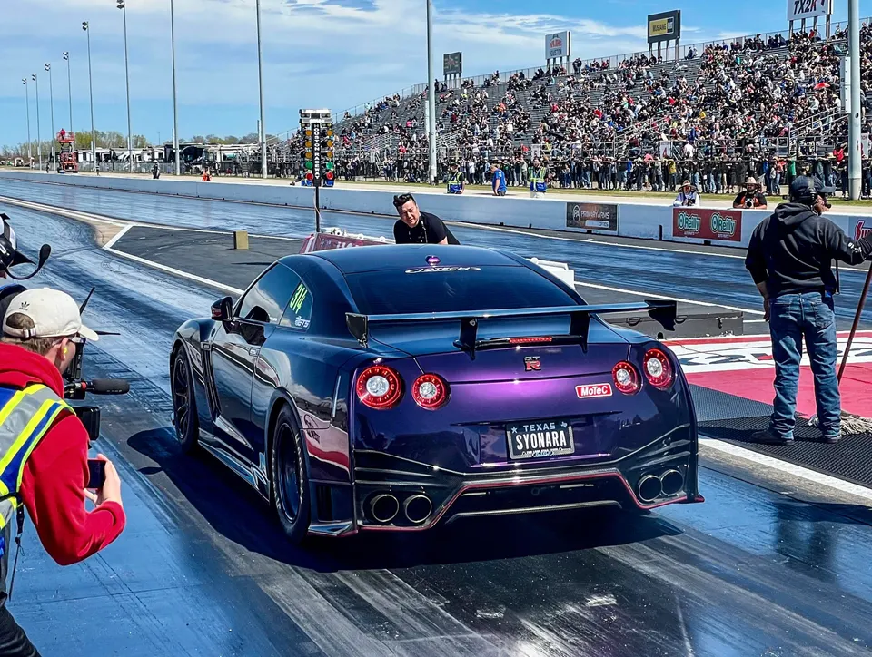 Purple Nissan GT-R with a large rear wing and custom exhaust at a drag strip, with spectators in the stands.