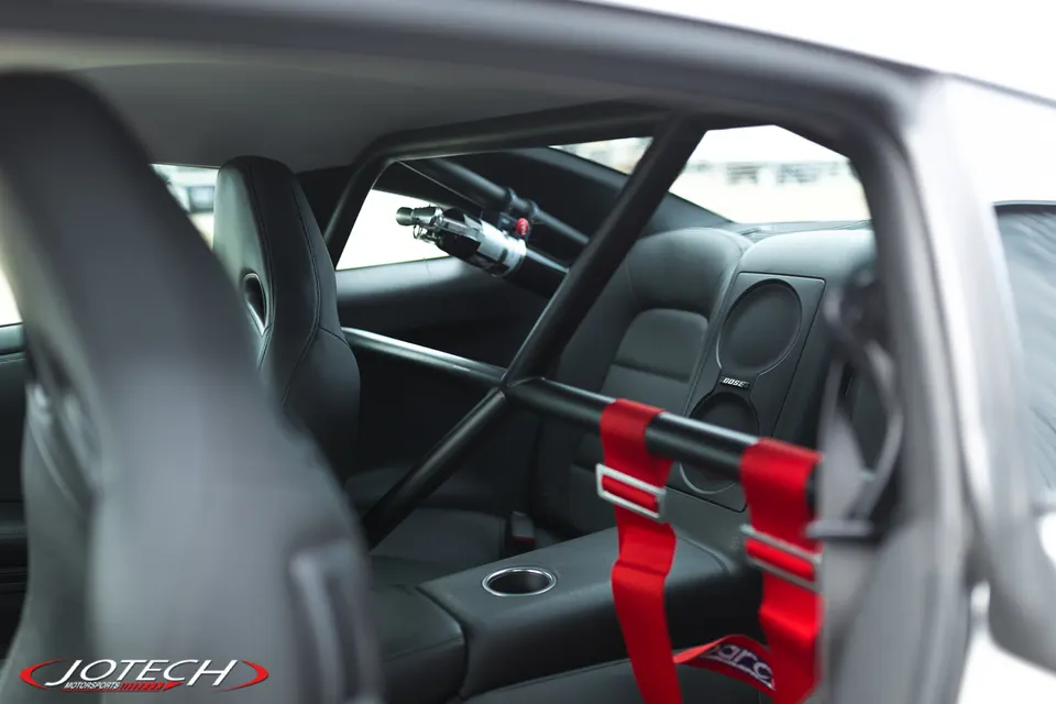 Interior view of a Nissan GT-R with a black roll cage, racing seats, red Sparco harnesses, and a fire extinguisher.