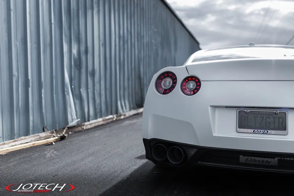 A white Nissan GT-R from the rear, showcasing its iconic taillights and GReddy exhaust tips, parked next to a corrugated metal wall.