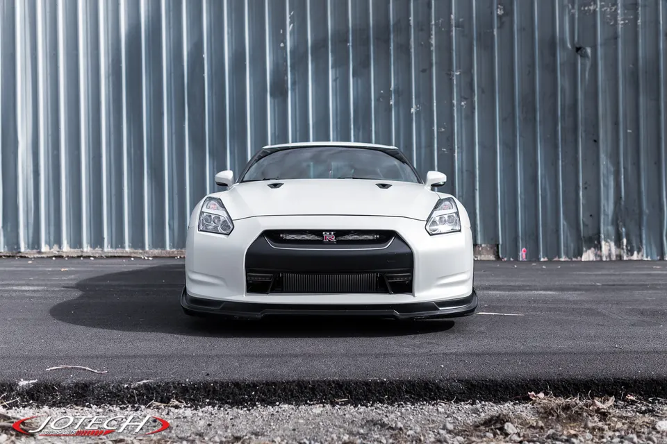 White Nissan GT-R with a front splitter parked in front of a corrugated metal wall, showcasing its aggressive front design.
