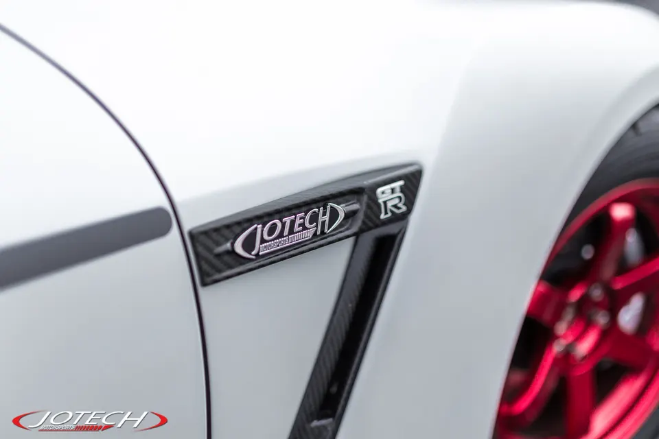 Close-up of a white Nissan GT-R fender with a Jotech Motorsports carbon fiber badge and a red wheel.