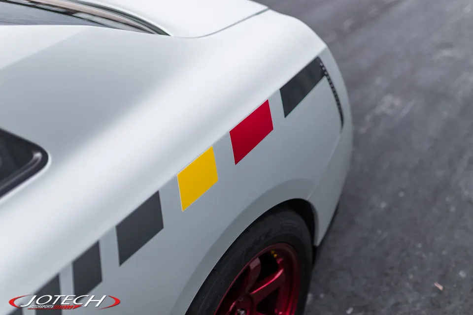 Close-up of a white car's rear quarter panel featuring custom black, yellow, and red stripe decals and a red wheel.
