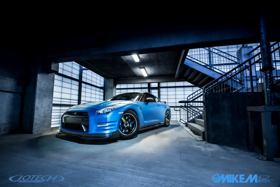A blue Nissan GT-R R35 with aftermarket wheels and a front splitter parked in a modern concrete parking garage.