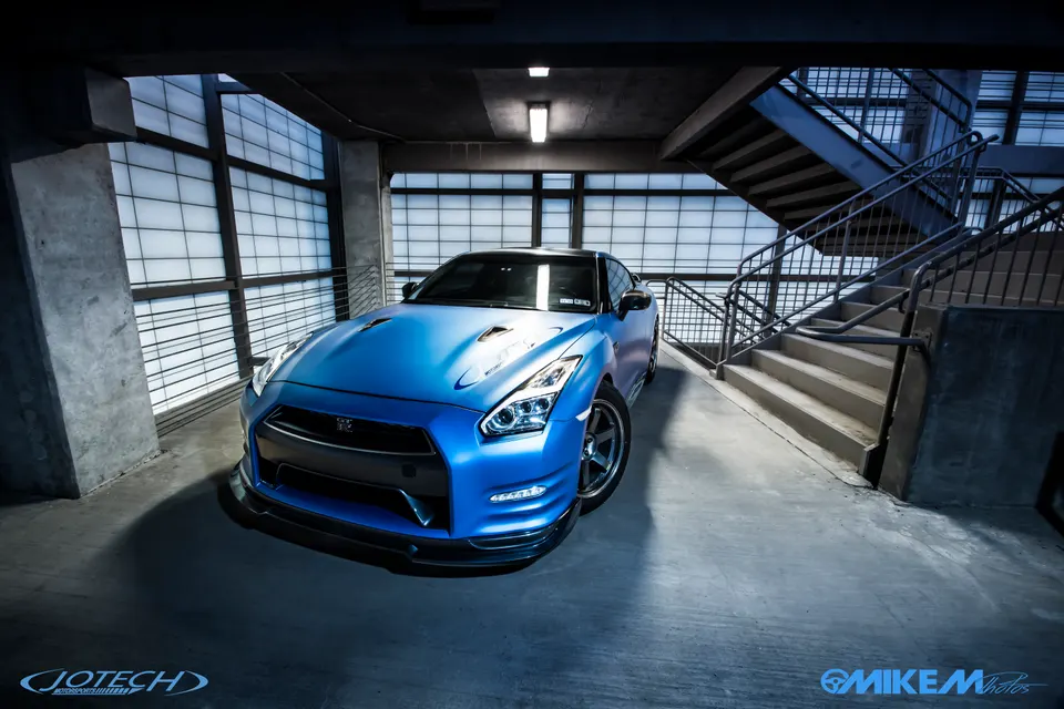 A blue Nissan GT-R R35 with aftermarket wheels and a front splitter parked in a modern parking garage.