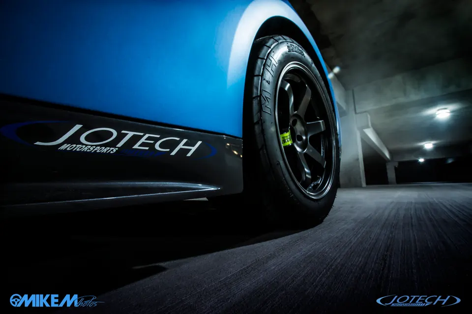Close-up of a blue car's wheel and Jotech Motorsports side skirt in a dimly lit parking garage.