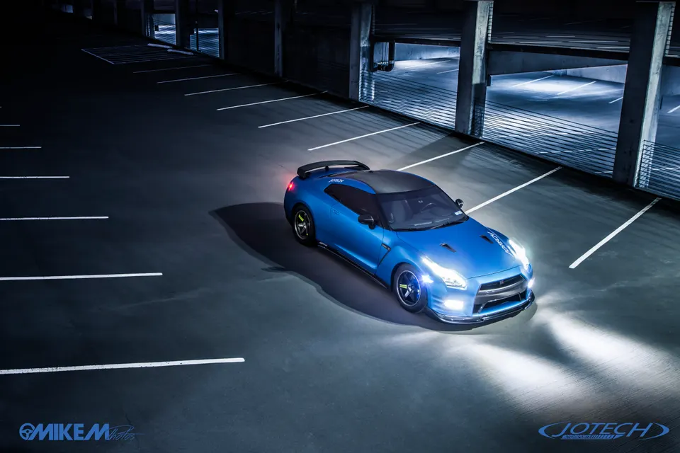 Blue Nissan GT-R with custom wheels and an aftermarket wing parked in a dimly lit parking garage at night.