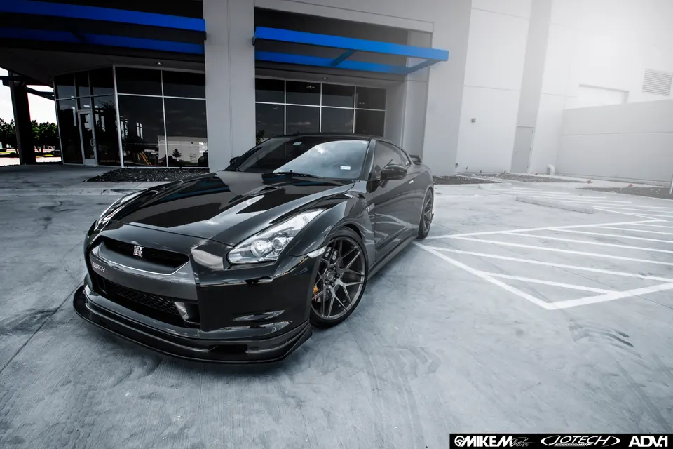 A sleek black Nissan GT-R with custom wheels and an aftermarket front splitter parked outside a modern building.