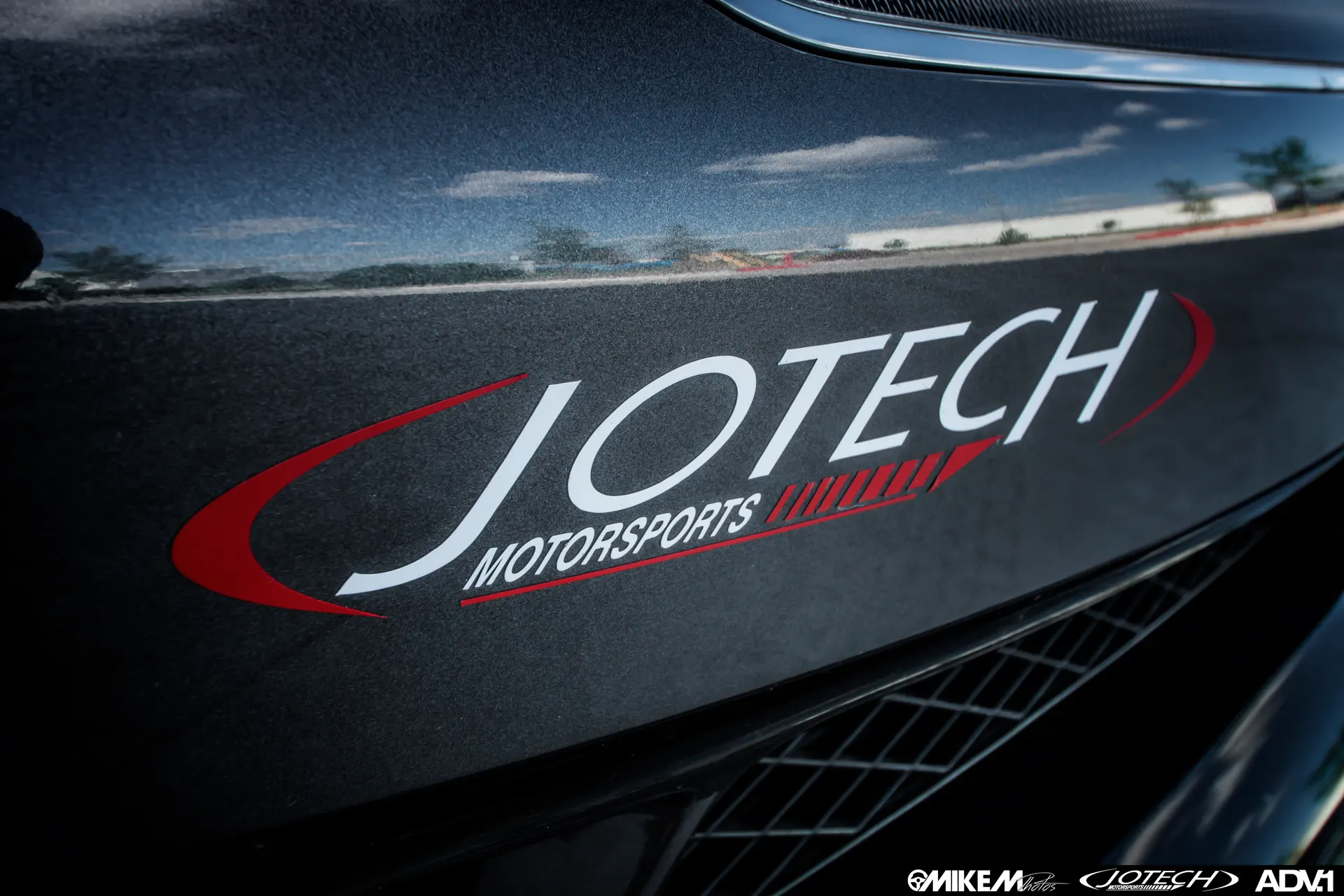 Close-up of a Jotech Motorsports decal on a dark grey car body, reflecting the sky.
