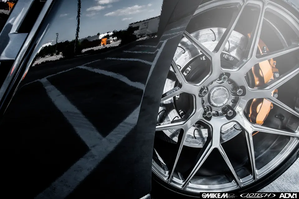 Close-up of a black car's custom multi-spoke wheel with orange performance brake calipers and a reflective body.