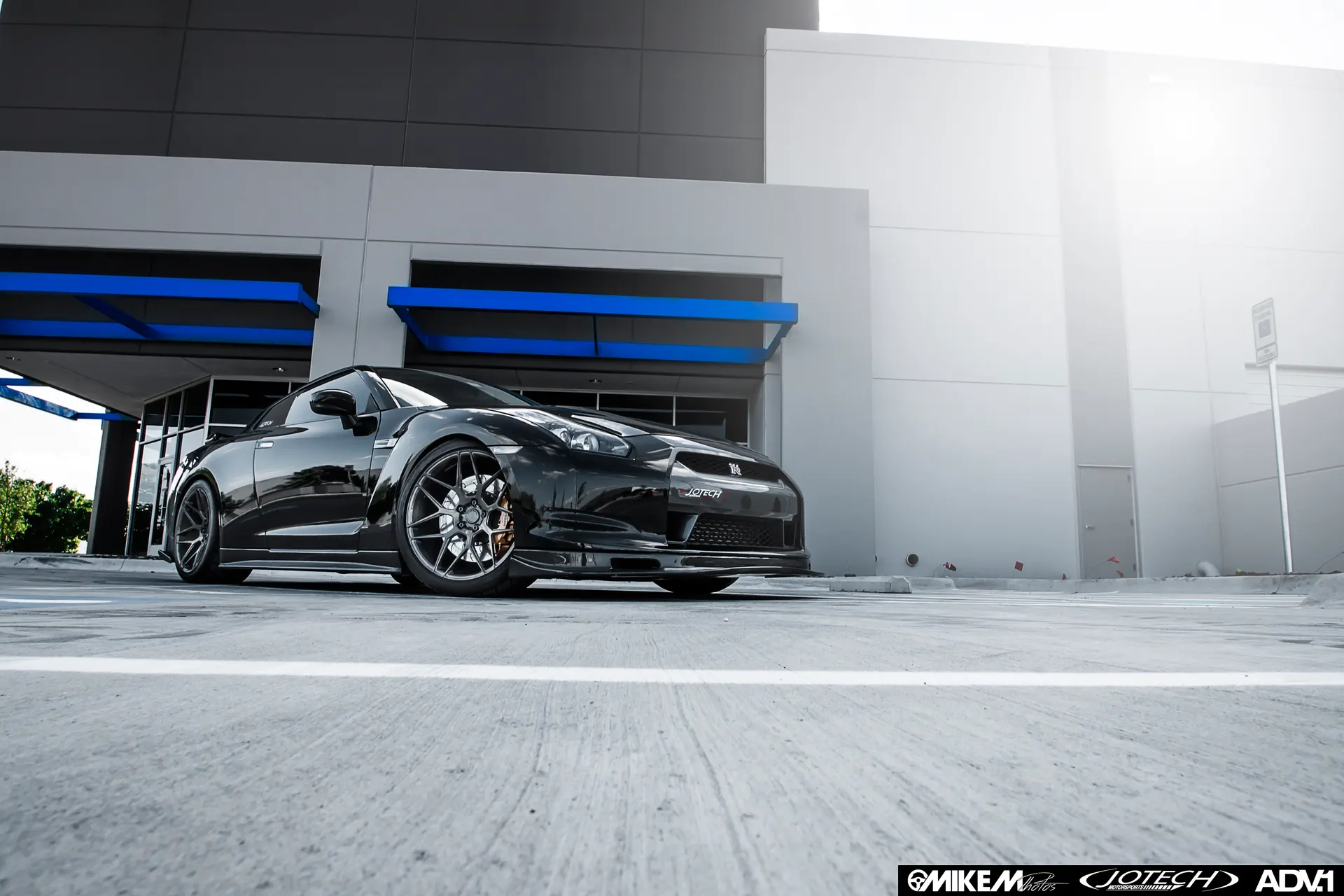 Black Nissan GT-R with custom ADV.1 wheels parked in front of a modern building with blue accents.