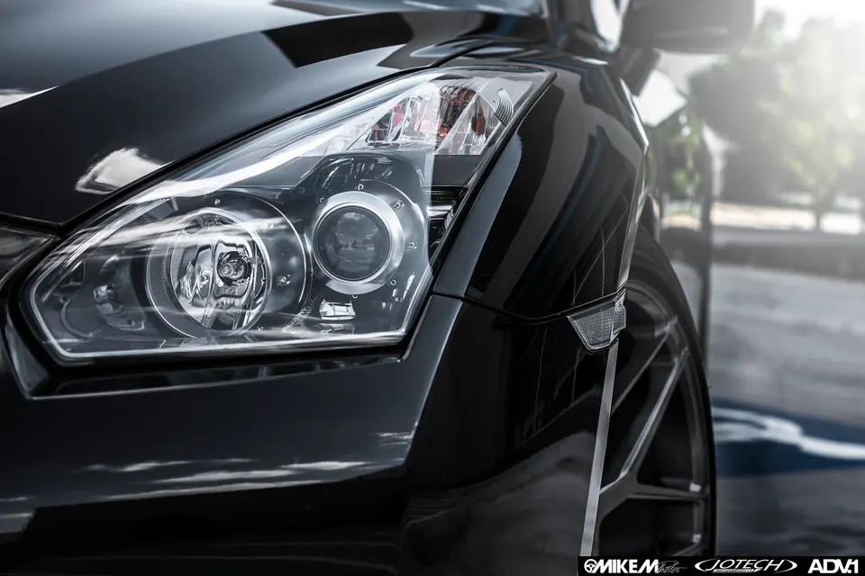 Close-up of a black Nissan GT-R's front headlight, fender, and custom ADV.1 wheel, showcasing exterior detail.