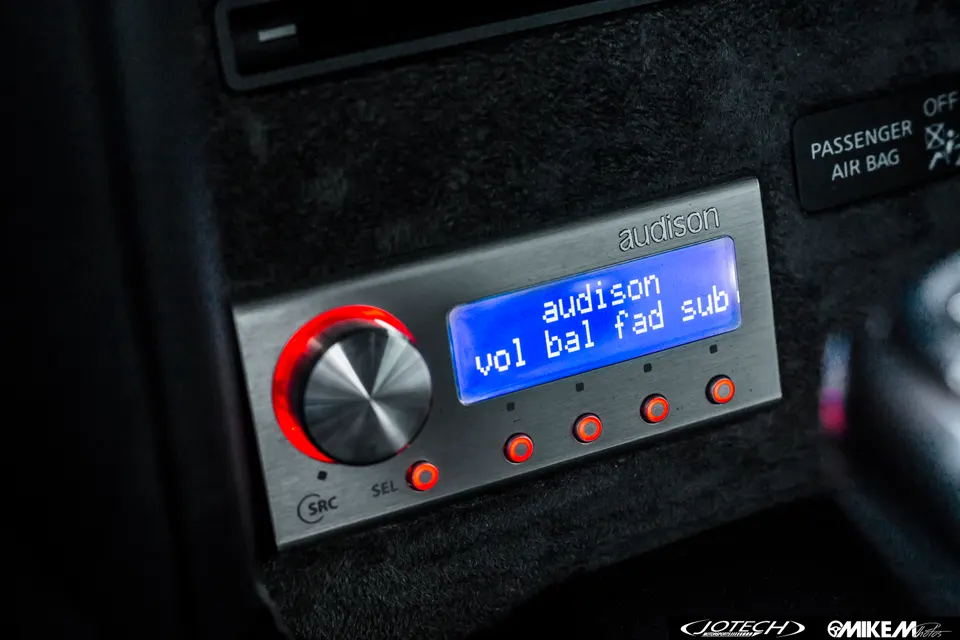 Close-up of an Audison car audio controller with a blue display and red backlit buttons in a car interior.