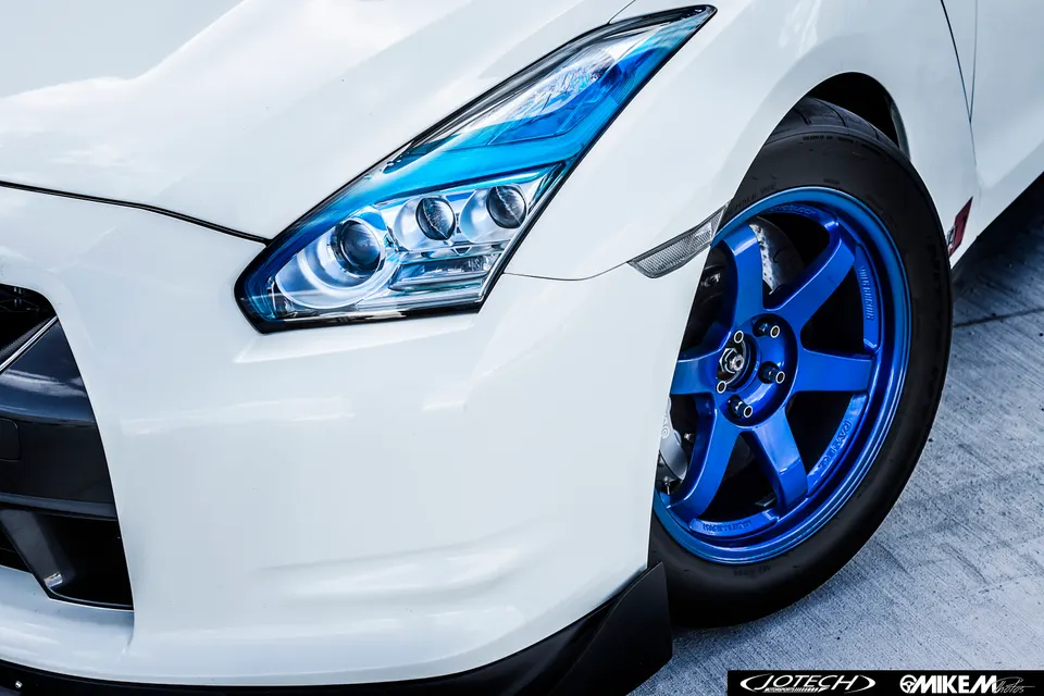 A close-up of a white Nissan GT-R's front quarter, showcasing its blue custom wheels and headlight.