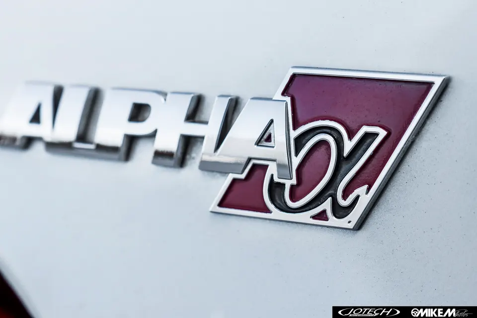 Close-up of a chrome Alpha badge with a red and black Alpha-X logo on a car's body panel.