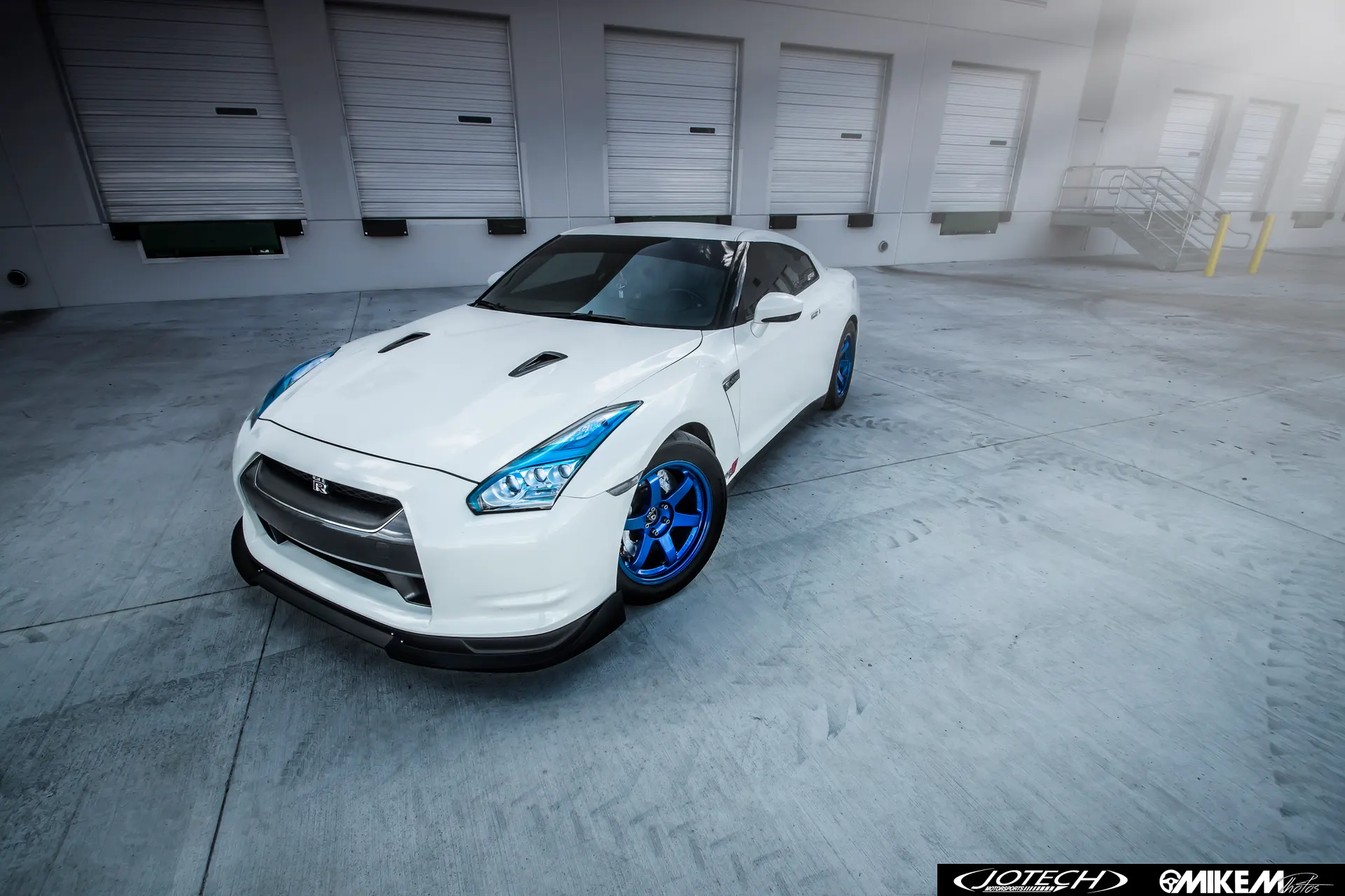 White Nissan GT-R with blue wheels and a front splitter parked in front of a warehouse loading dock.