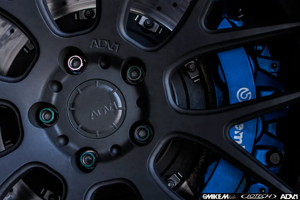 A close-up detail shot of a matte black ADV.1 wheel with blue Brembo brake calipers and iridescent lug nuts.