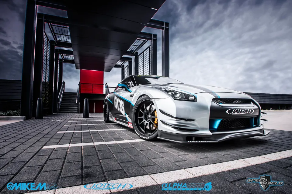 A silver and blue modified Nissan GT-R R35 with aftermarket wheels and aero kit parked in a modern architectural setting.