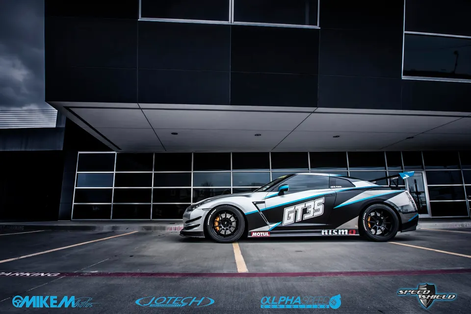 A silver and black Nissan GT-R with a "GT35" custom wrap and large rear wing parked in front of a modern building.