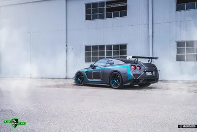 A customized Nissan GT-R with a matte grey and blue wrap, aftermarket wheels, and a large rear wing parked outside a building.