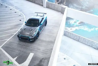 A modified Nissan GT-R with a large rear wing and custom livery parked on a multi-story structure overlooking the ocean.