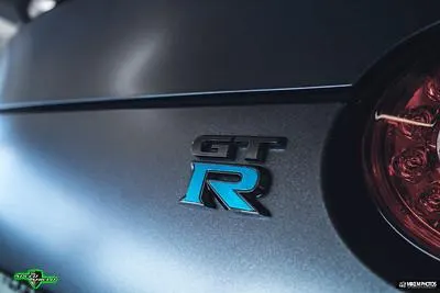 A close-up of the Nissan GT-R badge with a blue 'R' on a matte grey car, showing part of the red taillight.