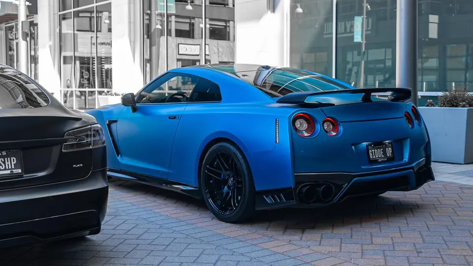 A matte blue Nissan GT-R with aftermarket black wheels and a rear wing parked in an urban setting.