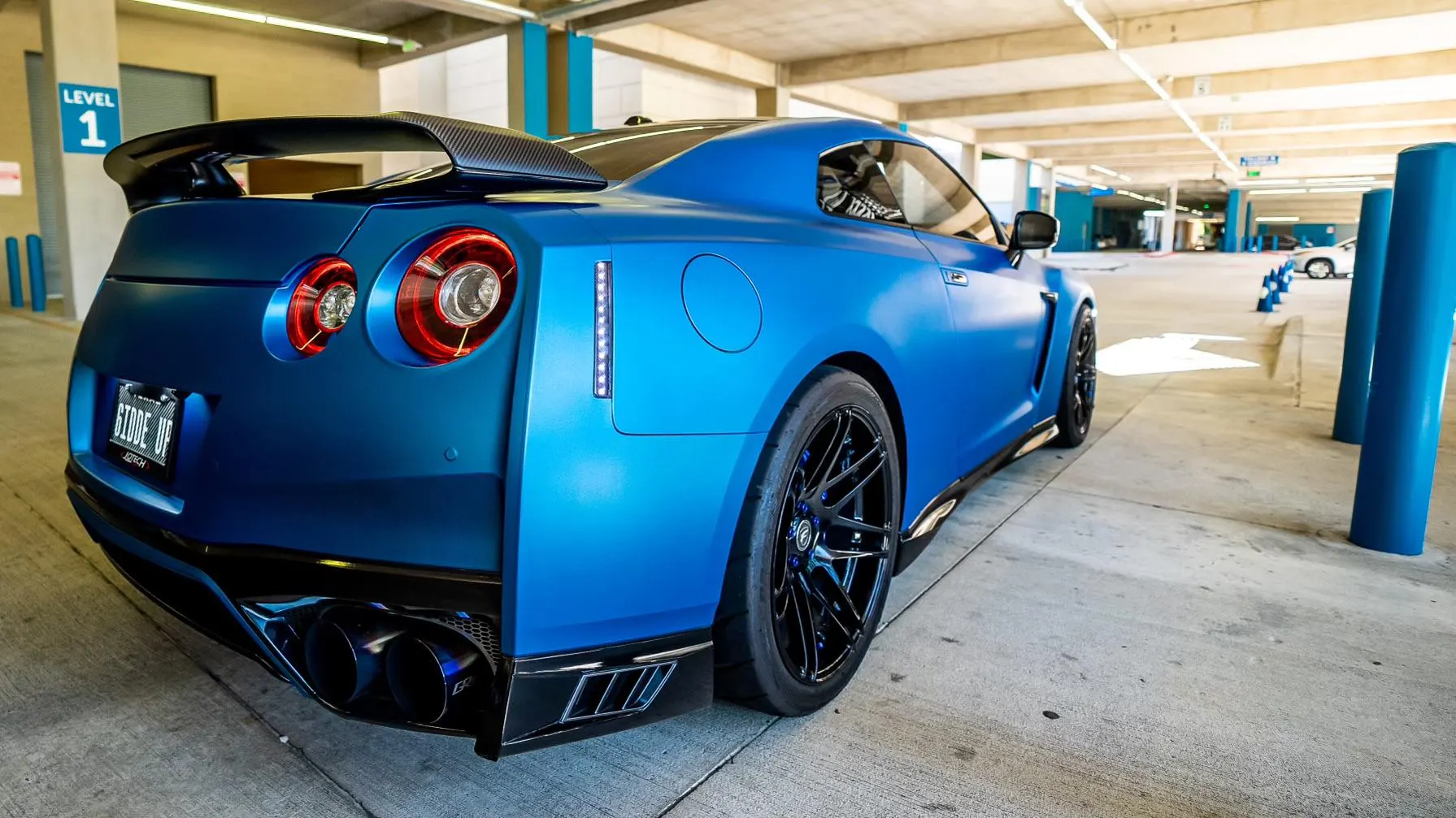 Matte blue Nissan GT-R R35 with a carbon fiber wing and aftermarket exhaust in a parking garage.
