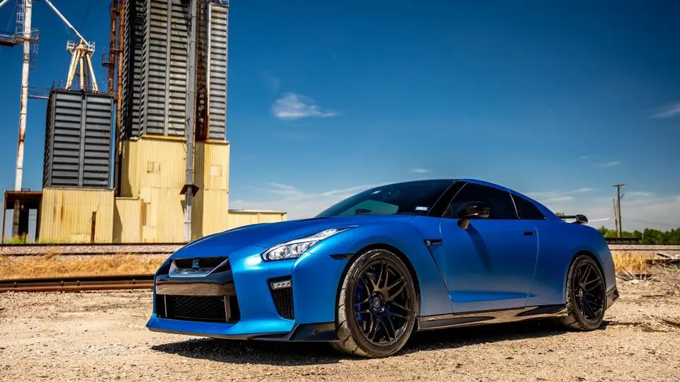 Blue Nissan GT-R R35 parked on a dirt road with industrial buildings in the background under a clear sky.