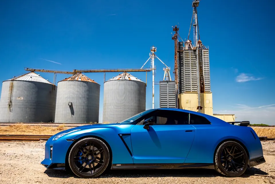 Blue Nissan GT-R sports car parked on a dirt road with large grain silos and industrial buildings in the background.