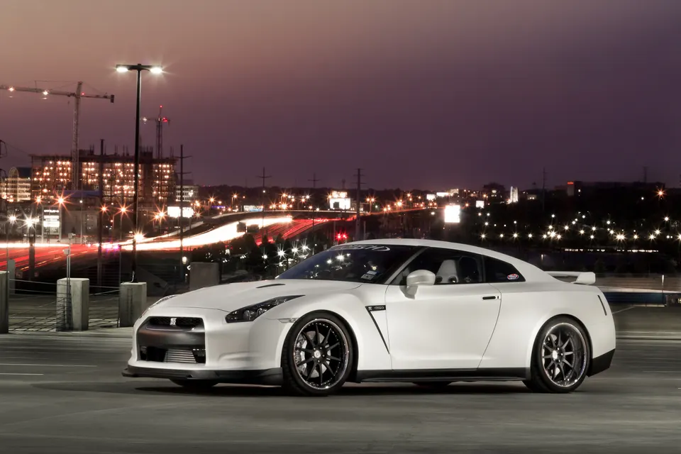 A white Nissan GT-R parked at night with city lights and light trails in the background, showcasing its sleek design.