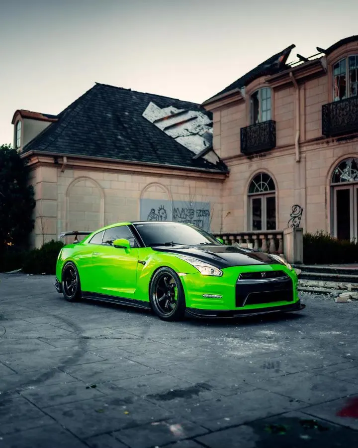 Bright green Nissan GT-R with a black hood and rear wing parked in front of a large, abandoned mansion.