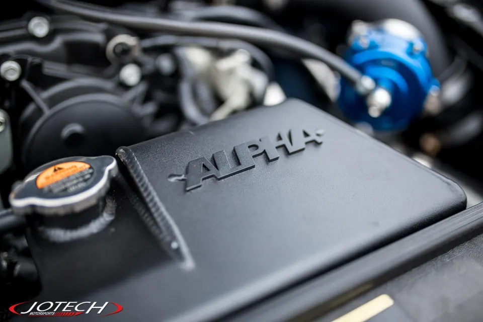 Close-up of a black Alpha Performance intercooler and a blue blow-off valve in a high-performance car's engine bay.