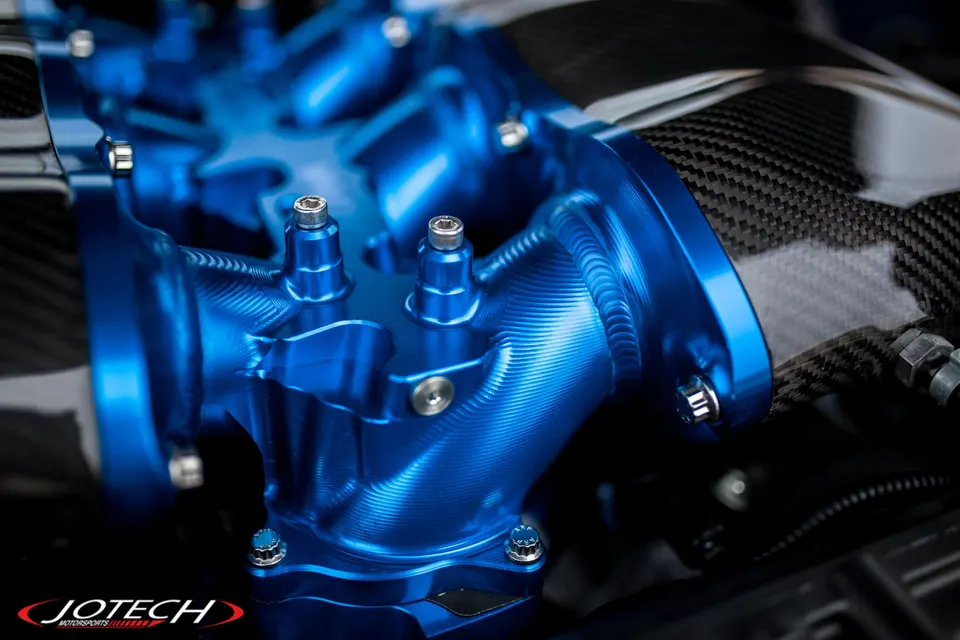 A close-up of a blue billet aluminum intake manifold with carbon fiber accents in an engine bay, featuring Jotech Motorsports branding.