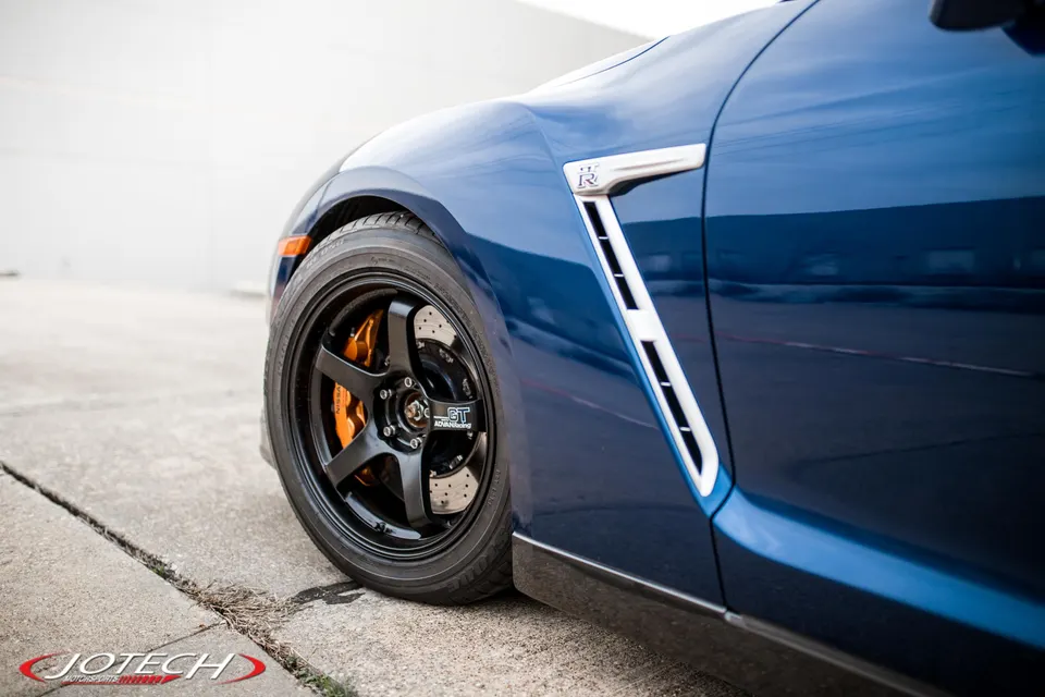 A close-up of a blue Nissan GT-R's front wheel, featuring black Volk TE37 wheels and orange brake calipers.