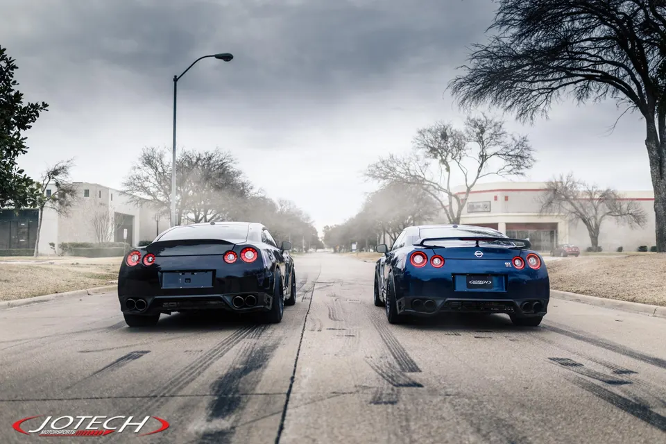 Black and blue Nissan GT-Rs parked on a street, showcasing their rear profiles with tire marks on the asphalt.