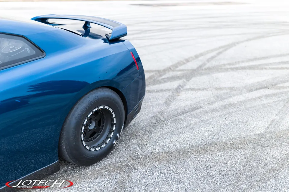 Rear quarter view of a blue Nissan GT-R with drag tires and beadlock wheels on an asphalt track with tire marks.