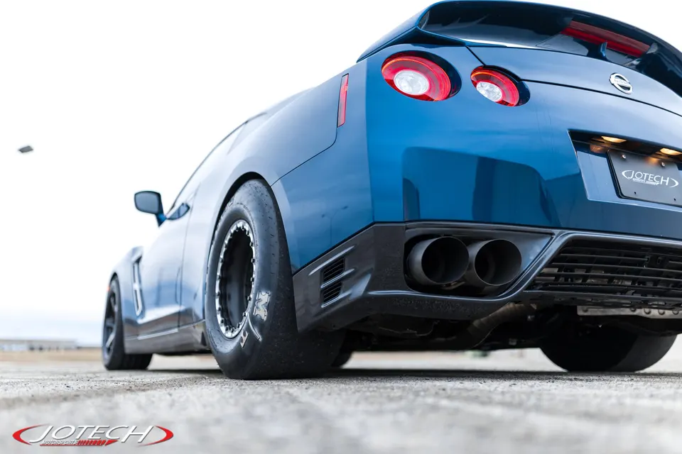 Blue Nissan GT-R with large drag slicks and aftermarket exhaust system, seen from a low rear angle on a paved surface.